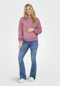 Felpa rosa chiaro con maniche lunghe e dettagli in pizzo, abbinata a jeans a palazzo blu e sneaker bianche. Il modello posa in modo casual.