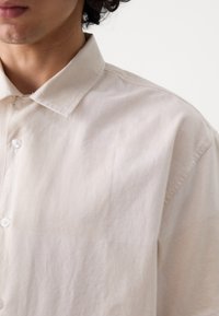 Chemise à manches courtes, beige clair, avec une texture douce, col pointu et fermetures à bouton, présentant une coupe décontractée et un design minimaliste.