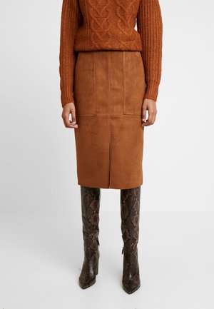 Kobieta ubrana w brązowy sweter w warkocze, pasującą brązową zamszową spódnicę midi z rozcięciem z przodu oraz brązowe, sięgające kolan buty ze skóry węża o spiczastych noskach.