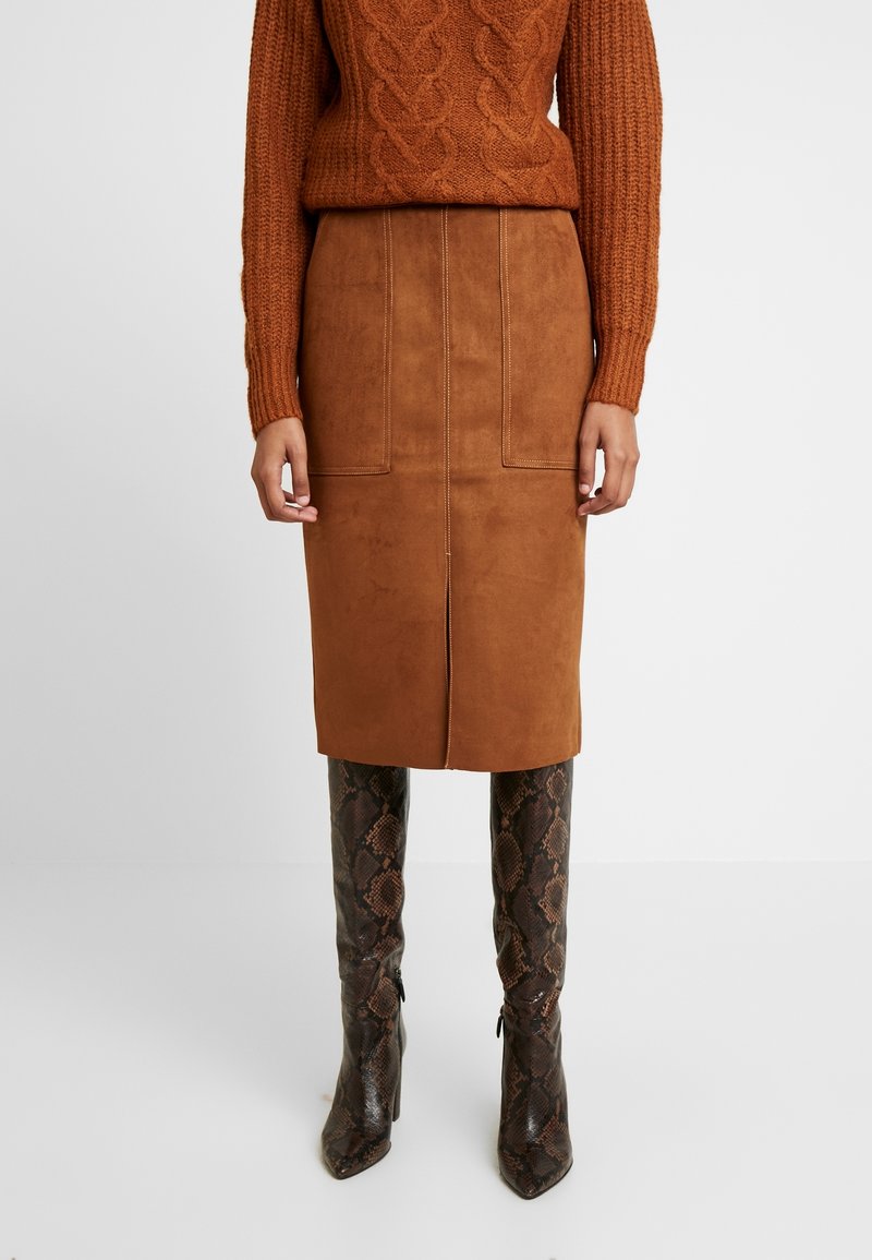 Femme portant un pull en maille torsadée marron, une jupe midi en daim marron assortie avec une fente à l'avant, et des bottes hauteur genou en cuir de serpent à bout pointu.