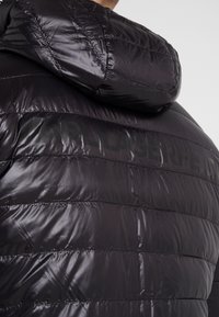 Schwarze glänzende gesteppte Jacke mit Kapuze, Rückansicht mit undeutlich eingeprägtem Markennamen über dem oberen Rücken.