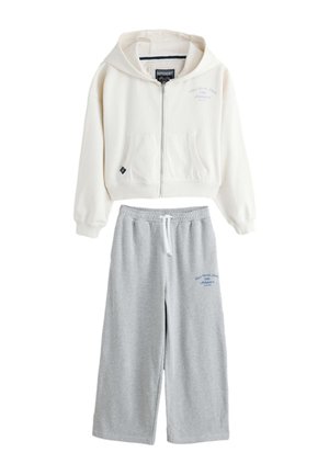 Hvid hoodie med lynlås og forlommer kombineret med grå vidde joggingbukser med snøre, begge med små blå tekstlogoer.