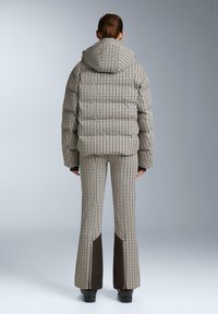 Prešívaná pufrová bunda s kapucňou v béžovo-hnedom károvanom vzore, spárovaná s nohavicami s rozšírenými nohavicami, ktoré majú tmavo hnedé akcenty.