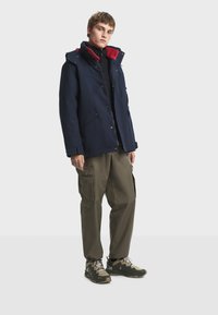 Giovane vestito con una giacca blu navy con rifiniture rosse, pantaloni cargo oliva e scarpe da trekking beige, in piedi contro uno sfondo chiaro e semplice.