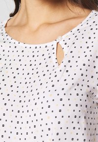 Top bianco con scollatura a chiave e chiusura a bottone, decorato con motivi a pois neri e gialli di diverse dimensioni. Tessuto morbido e liscio.