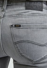 Jean en denim gris avec des coutures contrastantes, une étiquette sur la poche arrière et une finition légèrement texturée. La forme de la poche est arrondie avec peu d'ornements.