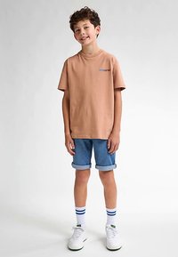 Vaaleanruskea lyhythihainen t-paita, jossa sininen teksti, yhdistettynä käännettyihin farkkushortseihin. Valkoiset tennarit ja sinivalkoraidalliset sukat täydentävät asun.