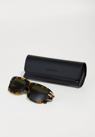 Occhiali da sole con montatura tartarugata e lenti scure accanto a una custodia in pelle nera Dolce & Gabbana su sfondo uniforme.