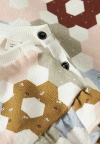 Pull en tricot présentant un motif hexagonal en rose, blanc, vert et marron avec de petites touches florales et un détail de bouton noir au col.