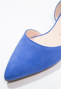 Chaussure plate bleue en daim à bout pointu avec côtés ouverts et doublure intérieure beige sur fond blanc.