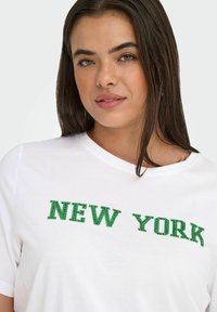 Moteris su ilgesniais tamsiais plaukais, vilkinti baltą marškinėlį su žaliais taškeliais, rašant „NEW YORK“ ant krūtinės.