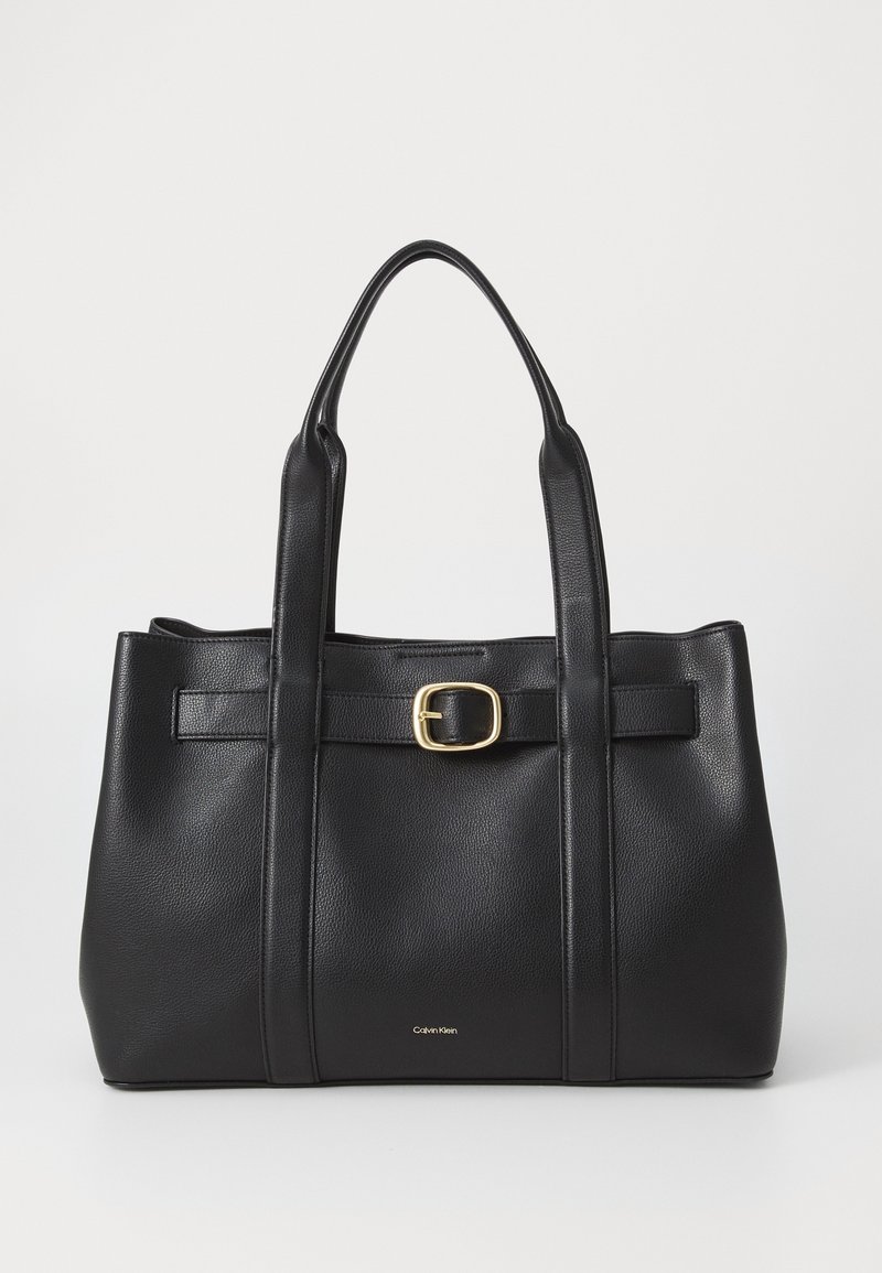 Sac cabas en cuir noir avec deux poignées, une ceinture avant avec une boucle dorée et un petit logo Calvin Klein près du bas au centre.