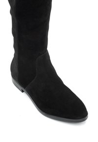 Botă din suede negru, până la genunchi, cu vârf ascuțit, detalii minime de cusătură și talpă plată. Textură netedă, design elegant.