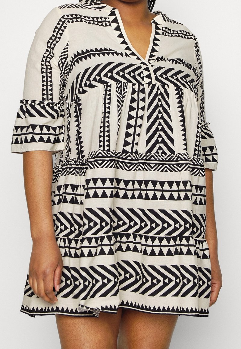 Femme portant une robe courte, ample, noire et blanche avec des motifs tribaux géométriques et des manches trois-quarts.