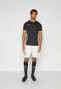 Camiseta atlética preta com detalhes dourados, calções brancos, meias negras até ao joelho com logótipo e chuteiras pretas, usados por um modelo masculino.