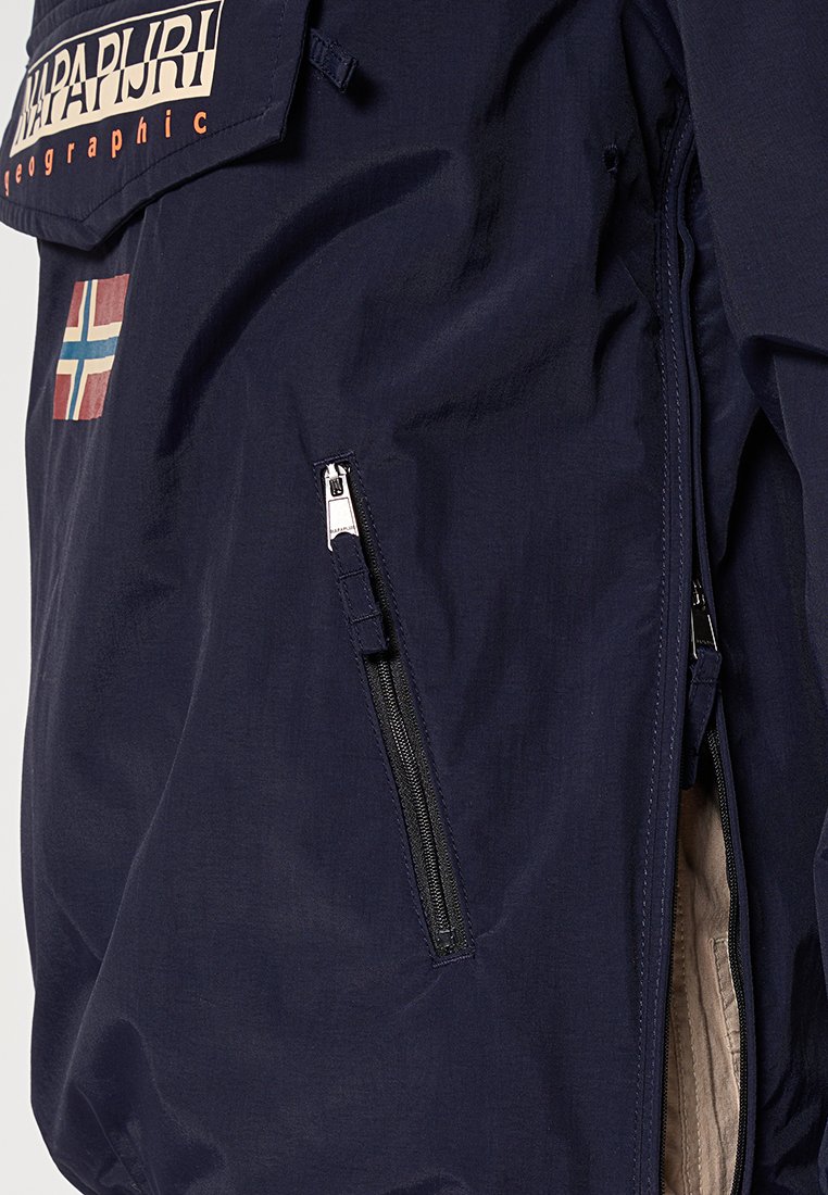 Section de la veste bleu marine montrant une poche zippée, un patch logo Napapijri et un petit patch drapeau norvégien sur le tissu.