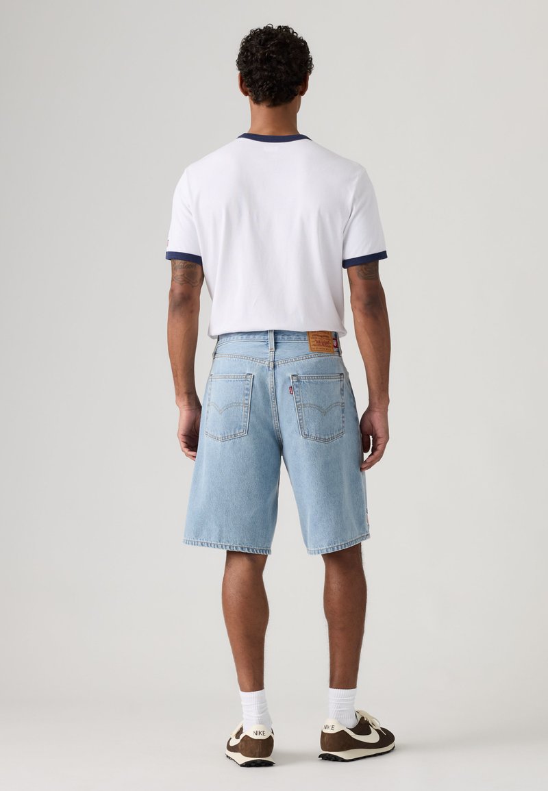 Mann trägt weißes T-Shirt mit marineblauem Rand, hellblaue Jeansshorts, weiße Socken und braune und weiße Nike-Sneakers, steht mit dem Rücken zur Kamera.