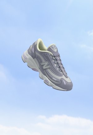 Une chaussure de sport grise et blanche unique avec le logo "N", flottant sur un fond de ciel bleu clair.