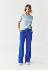 Light blue short-sleeve top paired with high-waisted blue trousers and white sneakers. Smooth fabric with a relaxed fit.
