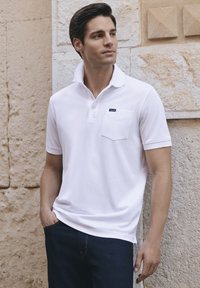 Polo shirt blanc en coton, avec un col en bouton, une poche poitrine, et un petit logo de marque. Porté avec un jean foncé.
