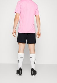Uomo in piedi di spalle, che indossa una maglietta sportiva rosa, pantaloni corti neri, calze bianche Adidas e scarpini neri Adidas Predator.
