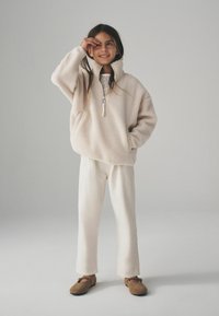 Ljusbeige fleecepullover med halvdragkedja och hög krage, tillsammans med krämfärgade vida byxor och bruna platta skor.