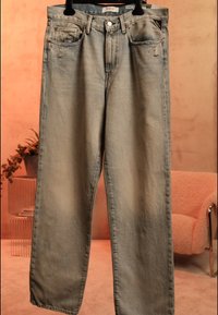 Jeans i lysvasket denim med en løs pasform, let slid ved lommerne, hængende på en sort bøjle mod en varmtonet indendørs baggrund.