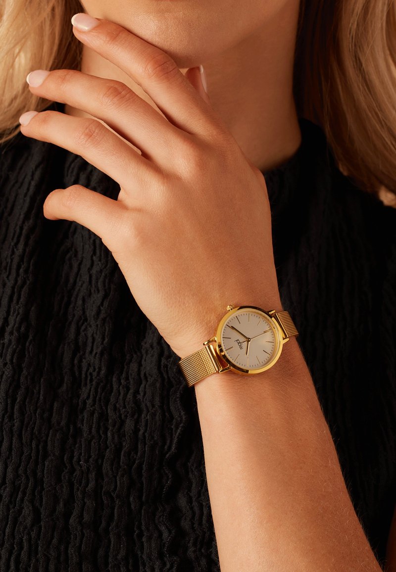Goldfarbene Uhr mit Mesh-Armband, runder silberner Zifferblatt und einfachen schwarzen Markierungen. Am Handgelenk vor einem strukturierten schwarzen Hintergrund getragen.