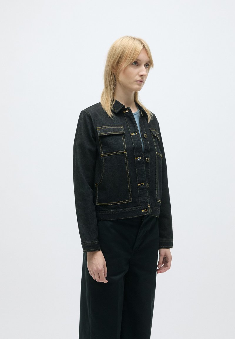 Chaqueta de denim negra con costuras amarillas, que presenta un cuello, cierre frontal con botones y dos grandes bolsillos en el pecho. La textura es suave y estructurada.