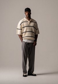 Polo din tricot cu dungi crem și negre, cu mâneci scurte, asortat cu pantaloni gri și pantofi negri. Modelul poartă o șapcă neagră.