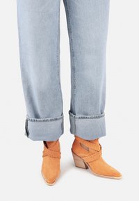 Bottines en daim orange avec un bout pointu et des accents tissés, associées à un jean bleu clair retroussé.