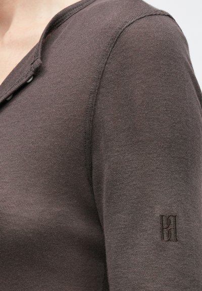 Közeli kép egy személyről, aki barna kötött hosszú ujjú felsőt visel, melyen a nyakrésznél két gomb található, és az ujjon egy kis hímzett logó látható.