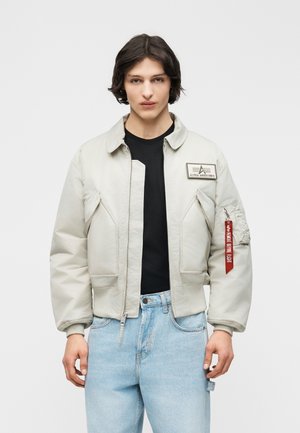 Νέος/-α με ανοιχτό μπεζ μπουφάν με φερμουάρ, που έχει το λογότυπο της Alpha Industries και κόκκινη ετικέτα «Remove Before Flight», μαύρο πουκάμισο και ανοιχτό μπλε τζιν.