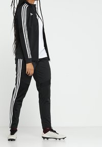Schwarzer sportlicher Trainingsanzug mit weißen Drei-Streifen-Akzenten, bestehend aus einer zip-festen Jacke und tapered Hosen, kombiniert mit weißen Fußballschuhen.