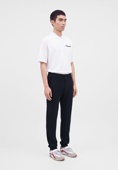 Jeune homme debout portant un polo blanc, un pantalon noir et des baskets grises avec des accents rouges sur un fond clair uni.
