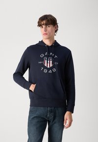 Sudadera de algodón azul marino con bolsillo canguro, capucha con cordón y un diseño gráfico en el frente que presenta un escudo y el año "1949".