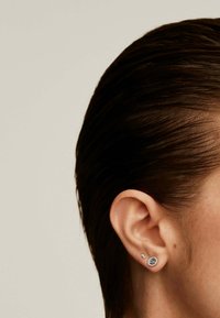 Pendiente de aro azul redondo con un halo de pequeñas piedras claras, acompañado de un pendiente en forma de estrella plateada más pequeño.