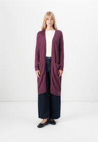 Vrouw staat tegen een witte achtergrond, gekleed in een lange paarse cardigan, een witte geribbelde top, wijde donkere spijkerbroek en zwarte ballet flats.