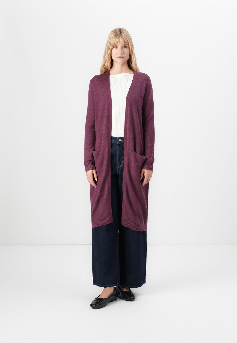 Vrouw staat tegen een witte achtergrond, gekleed in een lange paarse cardigan, een witte geribbelde top, wijde donkere spijkerbroek en zwarte ballet flats.
