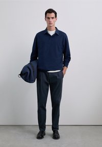 Camisa polo de manga larga azul marino con cuello, combinada con pantalones oscuros. Una bolsa azul marino está colgada de un brazo, y unos zapatos negros completan el conjunto.