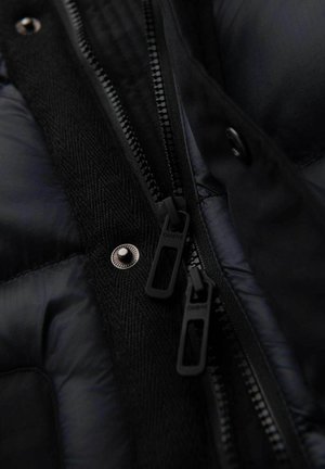 Primer plano de una chaqueta de plumas negra con tela suave, acentos texturizados y dobles cremalleras negras con marca. Presenta un detalle metálico de broche.