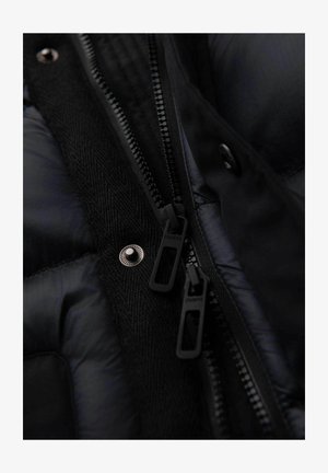 Primer plano de una chaqueta de plumas negra con tela suave, acentos texturizados y dobles cremalleras negras con marca. Presenta un detalle metálico de broche.