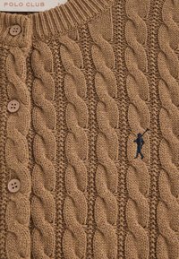 Brun kabelstickad kofta med fyra knappar, ribbad krage och ett broderat figuraccenter på framsidan. Texturerat ylletyg.