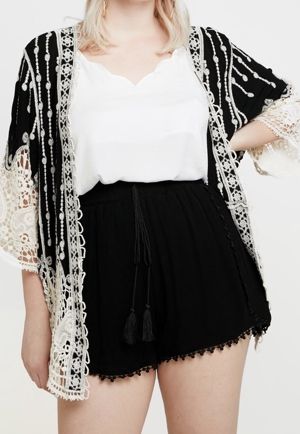 Kimono di pizzo nero con ricami bianchi intricati e maniche lunghe, indossato sopra una maglietta bianca e pantaloni corti neri con dettagli a frange.