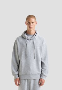 Felpa grigia chiara realizzata in tessuto morbido, con cappuccio regolabile, polsini a coste e una vestibilità comoda, indossata con pantaloni coordinati.
