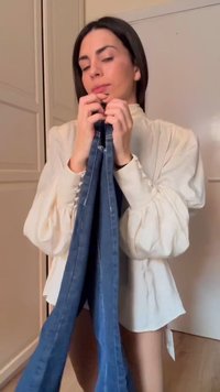 Γυναίκα με κρεμ μπλούζα κρατά μπλε τζιν από τη μέση, ετοιμάζεται να τα διπλώσει ή να τα κρεμάσει μέσα κοντά σε λευκές πόρτες ντουλάπας.