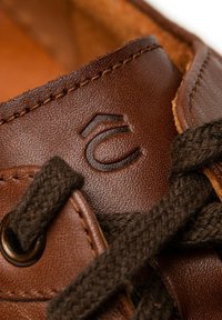 Brązowe skórzane buty z teksturowanym wykończeniem, z ciemnobrązowymi sznurowadłami, okrągłym metalowym oczkiem i wytłoczonym logo z boku.