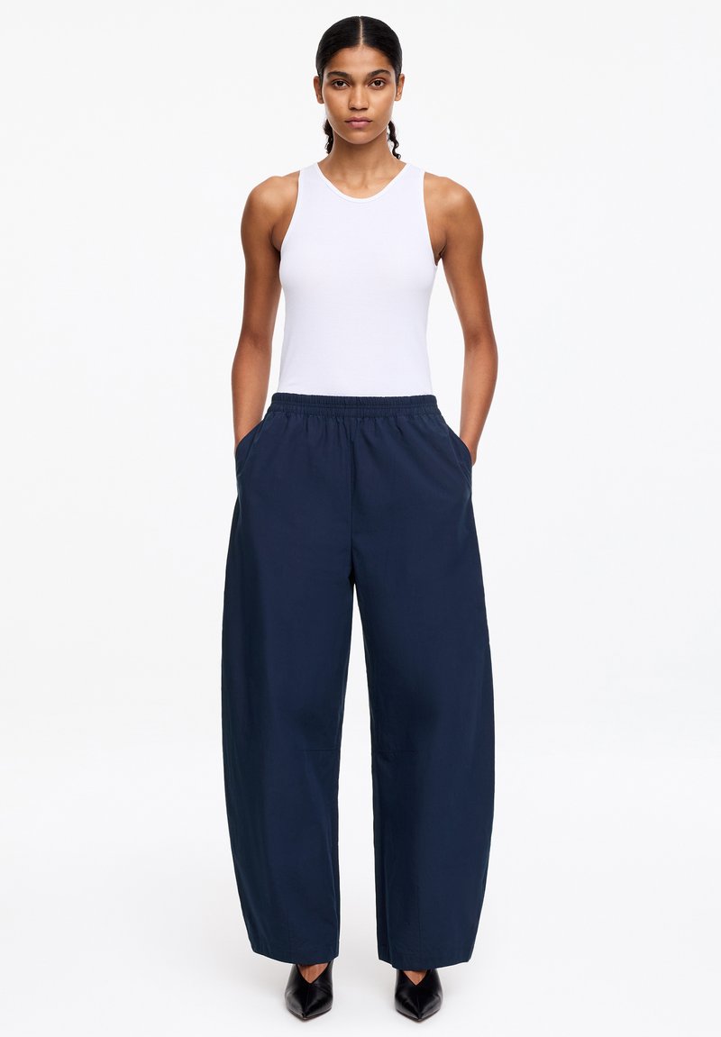 Femme portant un haut blanc sans manches, un pantalon large bleu marine avec les mains dans les poches, et des chaussures noires pointues sur un fond blanc.
