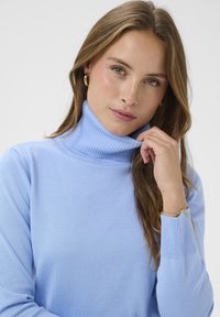 Világoskék garbó pulóver bordázott mintázattal és mandzsettákkal, testhezálló szabással. A modell a nyakát fogja, bemutatva a puha kötött anyagot.