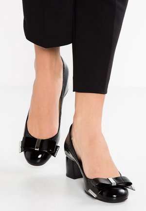 Femme portant des chaussures noires en cuir verni à talon bloc bas avec des nœuds décoratifs, associées à un pantalon noir raccourci sur un sol blanc.